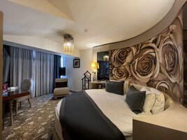 A hotel room with a large bed, a desk, a chair, and a view of the outdoors through a window with curtains.
