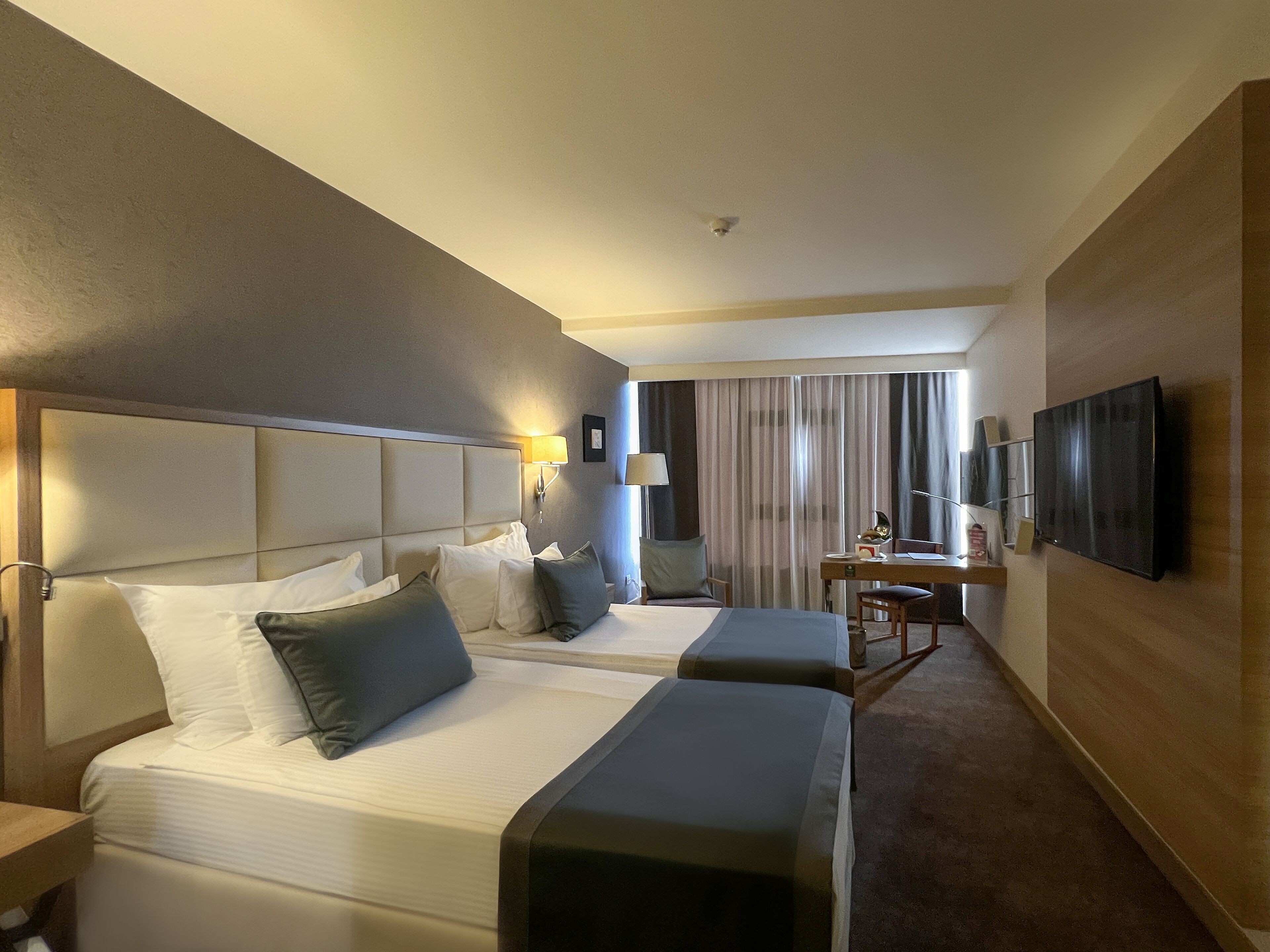 A hotel room with two beds, a desk, a TV, and a view of the cityscape.