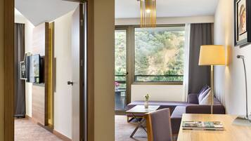 A modern hotel room with a wooden door, a sofa, a dining table, and a large window offering a view of greenery.
