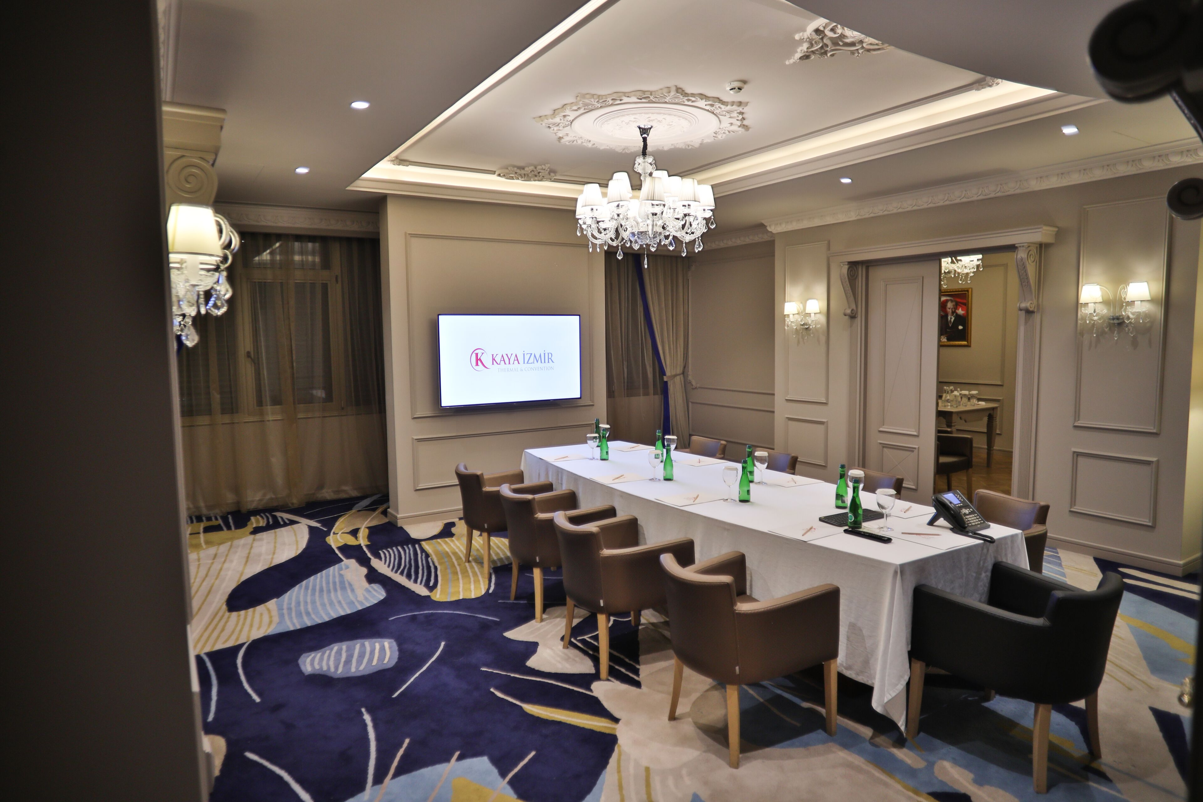 A conference room with a long table, chairs, a chandelier, and a screen displaying "KAYA IZMIR".