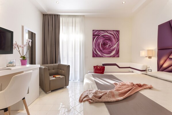 Una habitación de hotel moderna con una cama, un escritorio, una silla, un sofá, un televisor y un ventanal con cortinas.