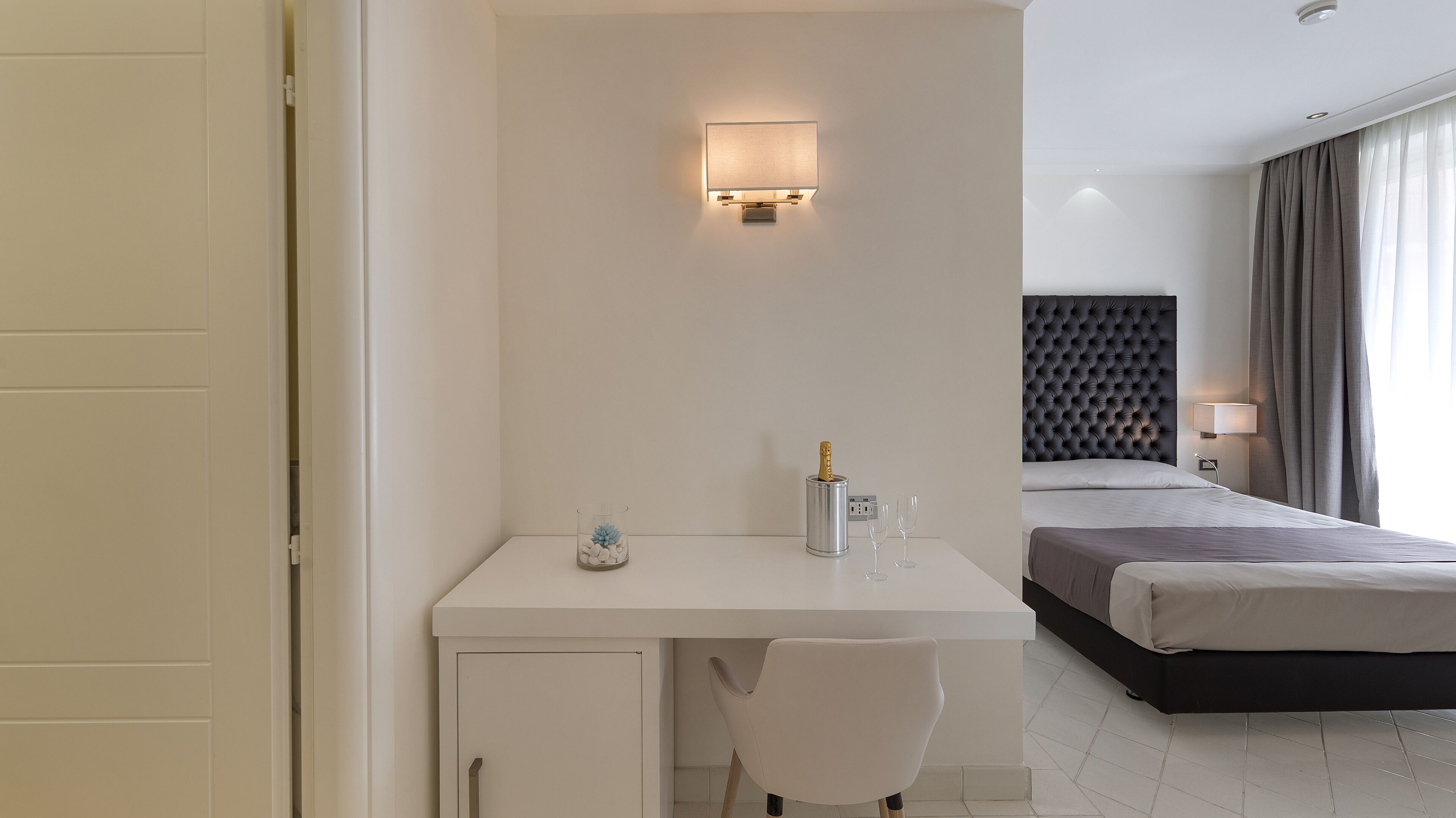 A hotel room with a white desk, a chair, a bed with a dark headboard, and a bedside table with a lamp.