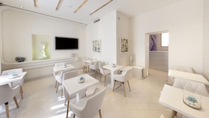 A modern, well-lit dining area with white tables and chairs, a flat-screen TV mounted on the wall, and artwork on the walls. - Palazzo Montefusco Relais (Sorrento)