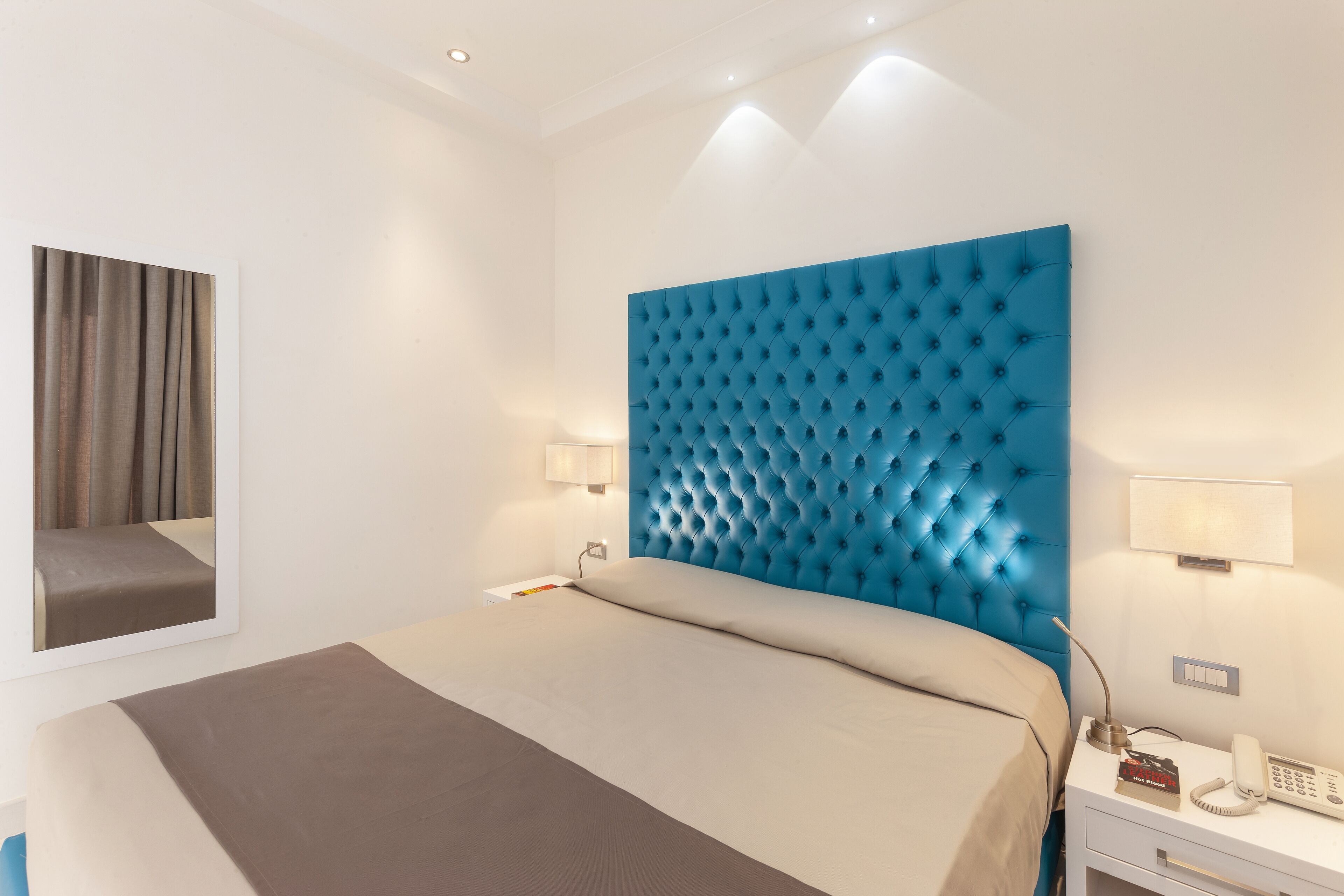Habitación de hotel con una cama grande, un cabecero azul con botoneras, lámparas de noche, una mesita con un teléfono y un espejo en la pared.