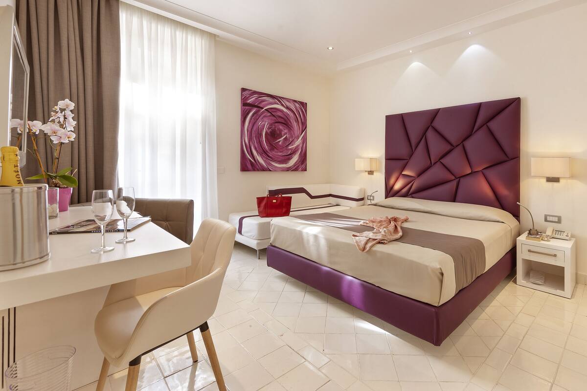 Ein modernes Hotelzimmer mit einem großen Bett, einem Schreibtisch mit zwei Stühlen, einem kleinen Tisch mit einer Vase und Gläsern, einer roten Handtasche und einem an der Wand befestigten Telefon.