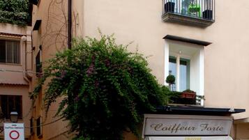Une rue animée, avec des piétons, un bâtiment sur lequel on peut lire l’inscription « Coiffeur Forte », et un arbre aux fleurs violettes.