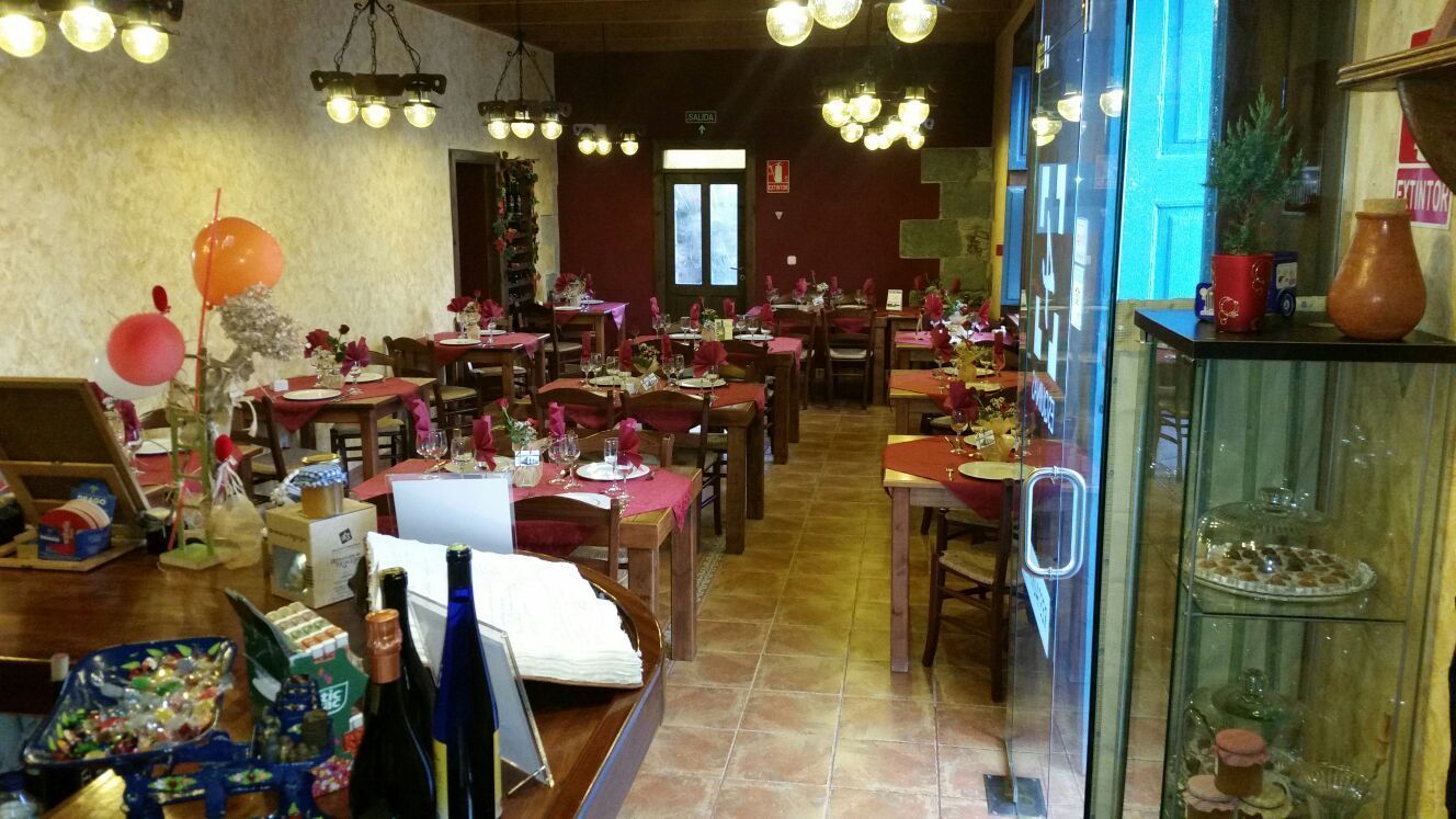 Un restaurante con varias mesas dispuestas, cubiertas con manteles rojos, velas y flores. Se observan lámparas colgantes y una vitrina con cristalería a la derecha.