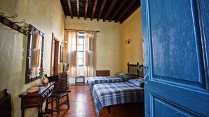 Quarto com piso de madeira, cama com colcha xadrez, uma cadeira, cômoda, espelho e janela com cortinas.