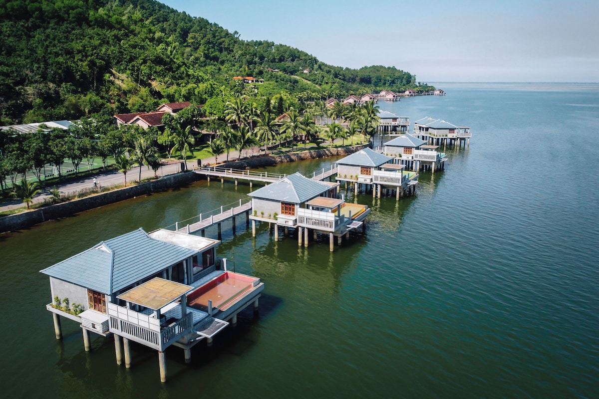 Overwater bungalows with a view of the sea and a forested hillside.