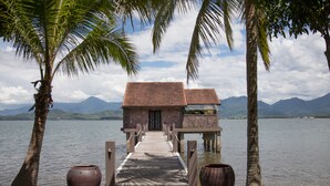 A wooden dock leading to a stone building with a tiled roof, surrounded by palm trees and a lake with mountains in the background. - Vedana Lagoon Resort and Spa (Hue)
