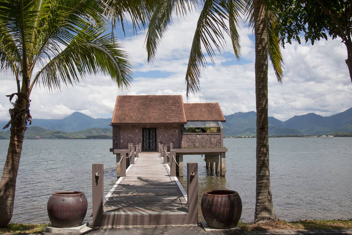 Eine Holzanlegestelle führt zu einem Steinhaus mit Ziegeldach, umgeben von Palmen und einem See mit Bergen im Hintergrund.