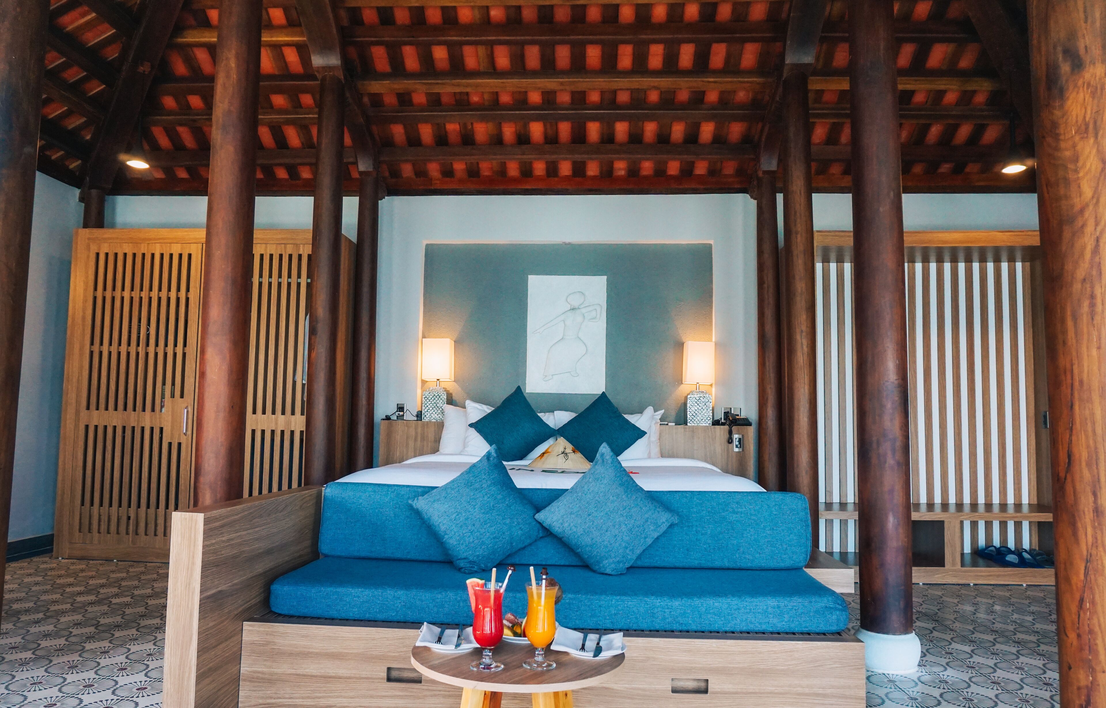 A hotel room with a bed, blue cushions, a wooden headboard, a nightstand with a lamp, and a tray with drinks.