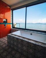 Un baño con una bañera grande, baldosas estampadas en rojo y blanco, y vista a una masa de agua a través de un ventanal.