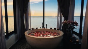 A large bathtub filled with rose petals, a vase with flowers, and candles, with a view of the sea through large windows. - Vedana Lagoon Resort and Spa (Hue)