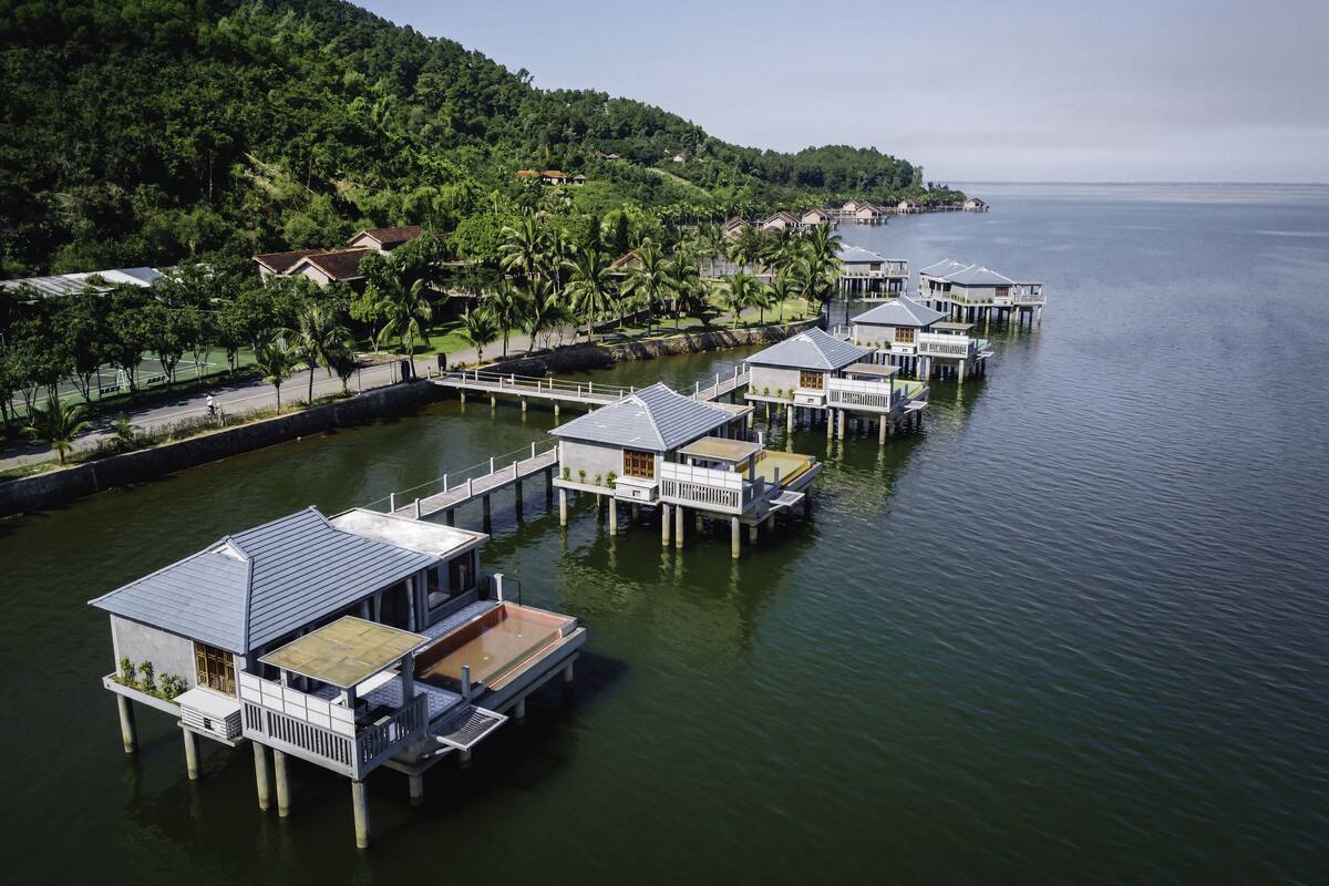 Bungalows auf Stelzen mit Blick auf das Meer und einen bewaldeten Hügel.