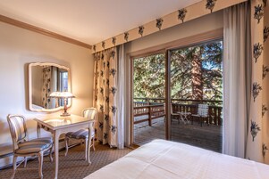 A hotel room with a bed, a desk with a lamp, two striped chairs, a large mirror, and a view of an outdoor deck with a wooden railing and chairs.
