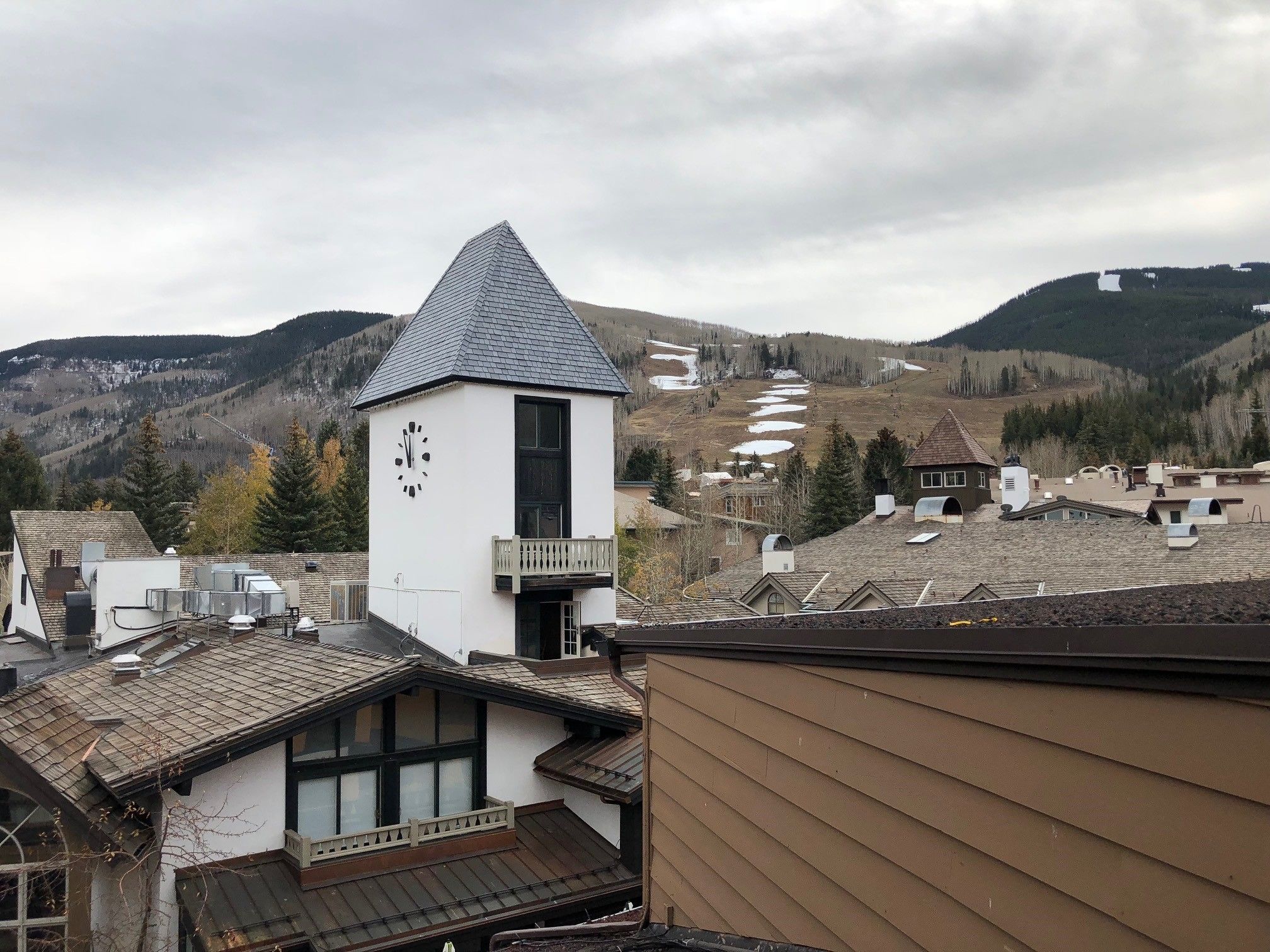 A small white building with a clock, surrounded by houses with sloped roofs, set against a backdrop of mountains and a cloudy sky.