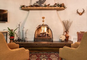 Una habitación acogedora con chimenea, dos sillones, un tapete rojo estampado y una estantería con piñas y follaje.