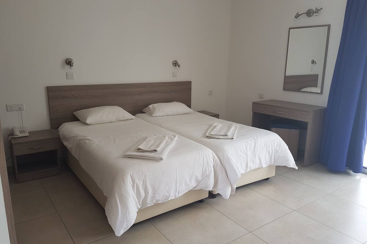 Una camera d'albergo con due letti, un comodino con una lampada, uno specchio e una finestra con tende.
