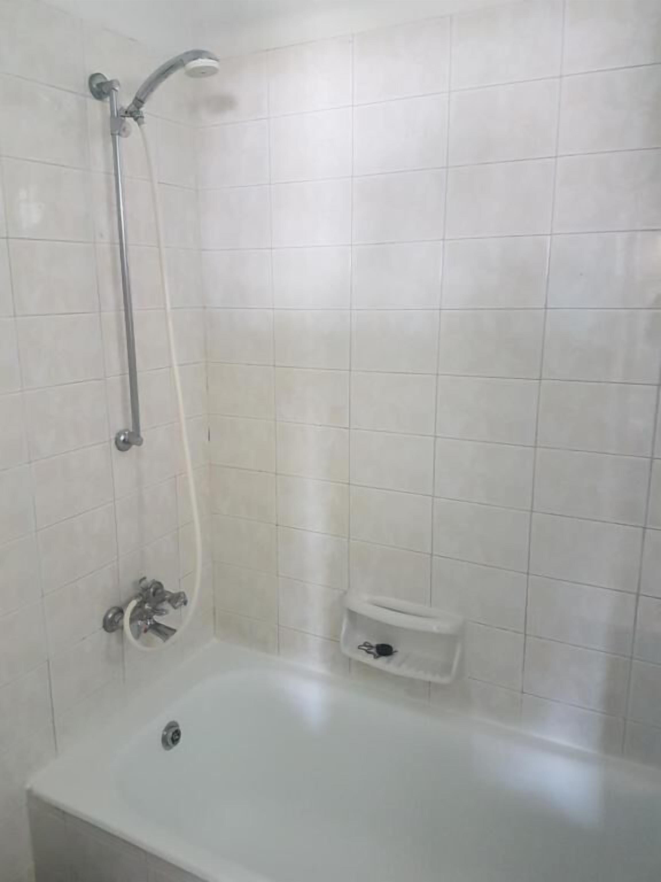 A white tiled shower area with a handheld showerhead, a handheld shower attachment, and a small white shelf.