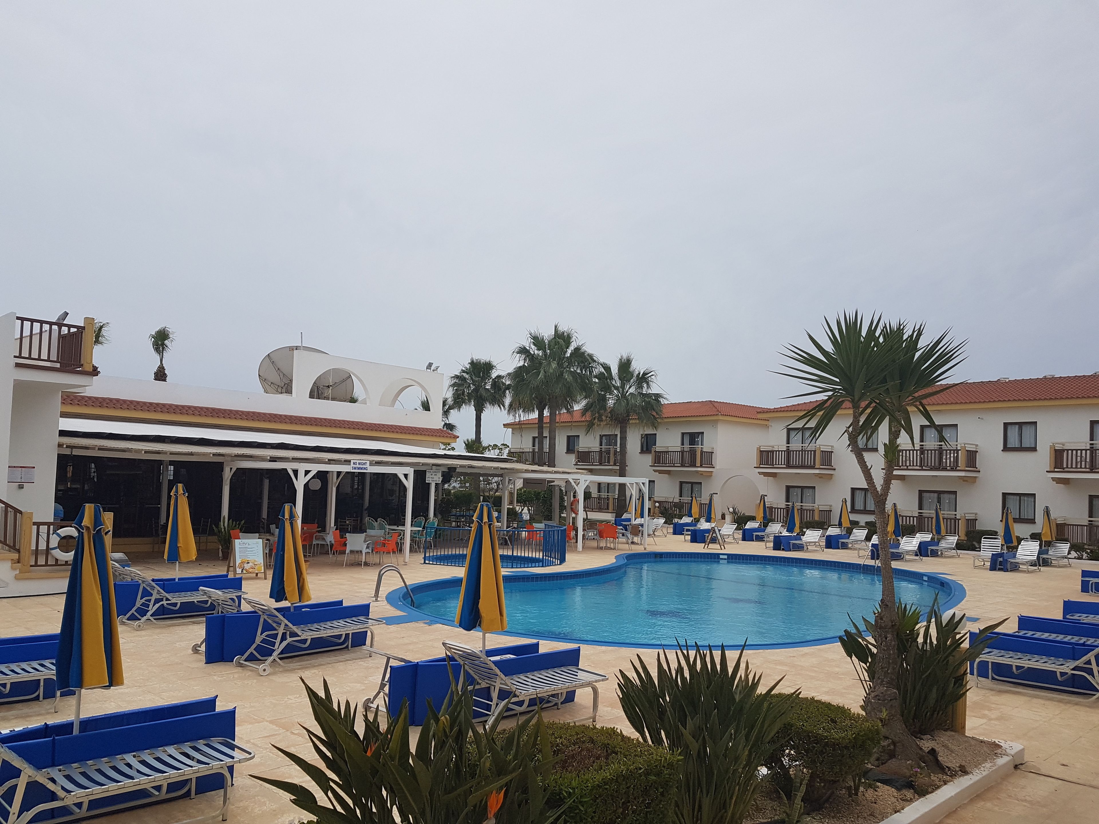 Uma área de piscina com espreguiçadeiras azuis e amarelas, piscina e um edifício com varandas e palmeiras.