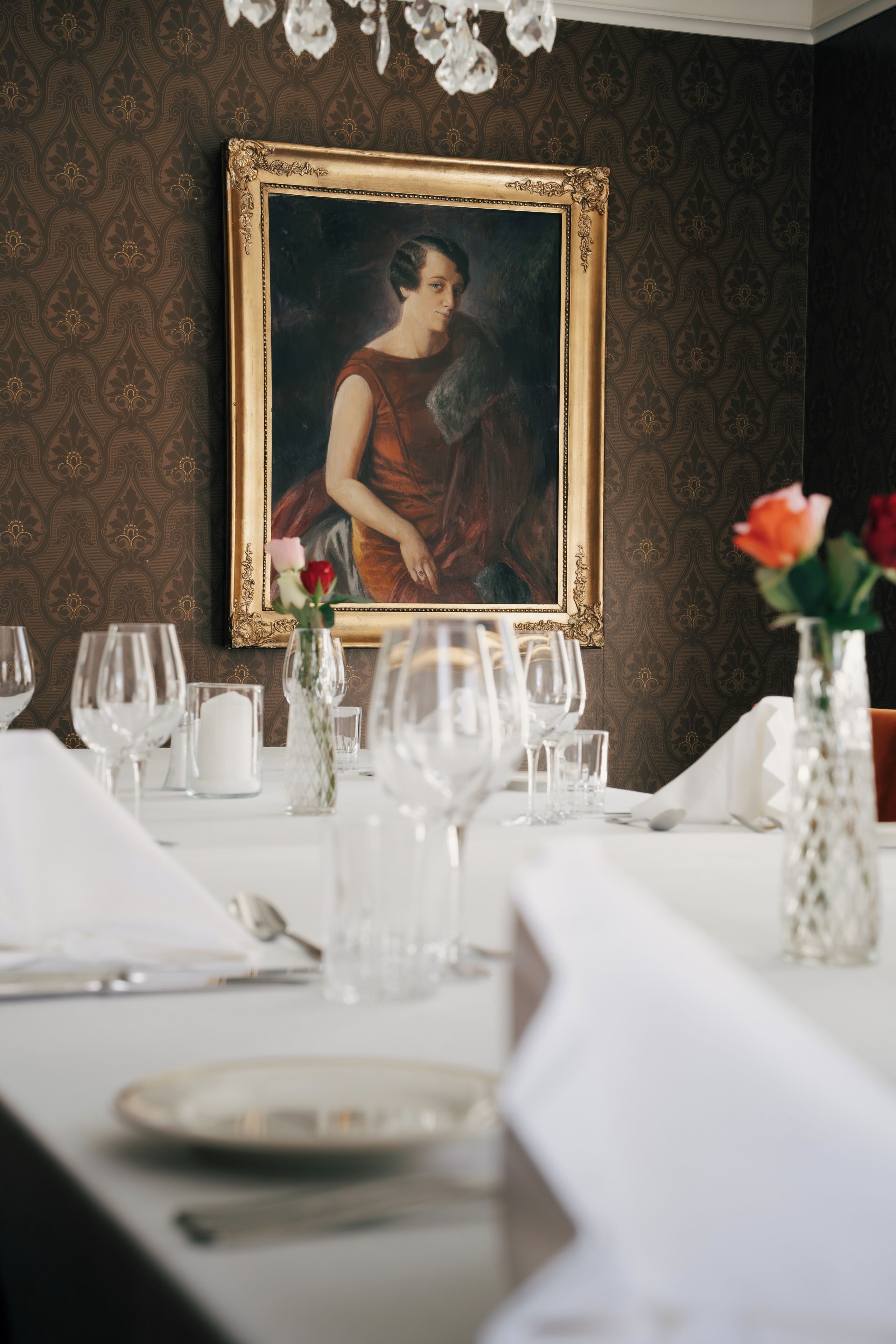 A dining table set with glassware and a vase with flowers, a framed portrait on the wall, and a patterned wallpaper background.
