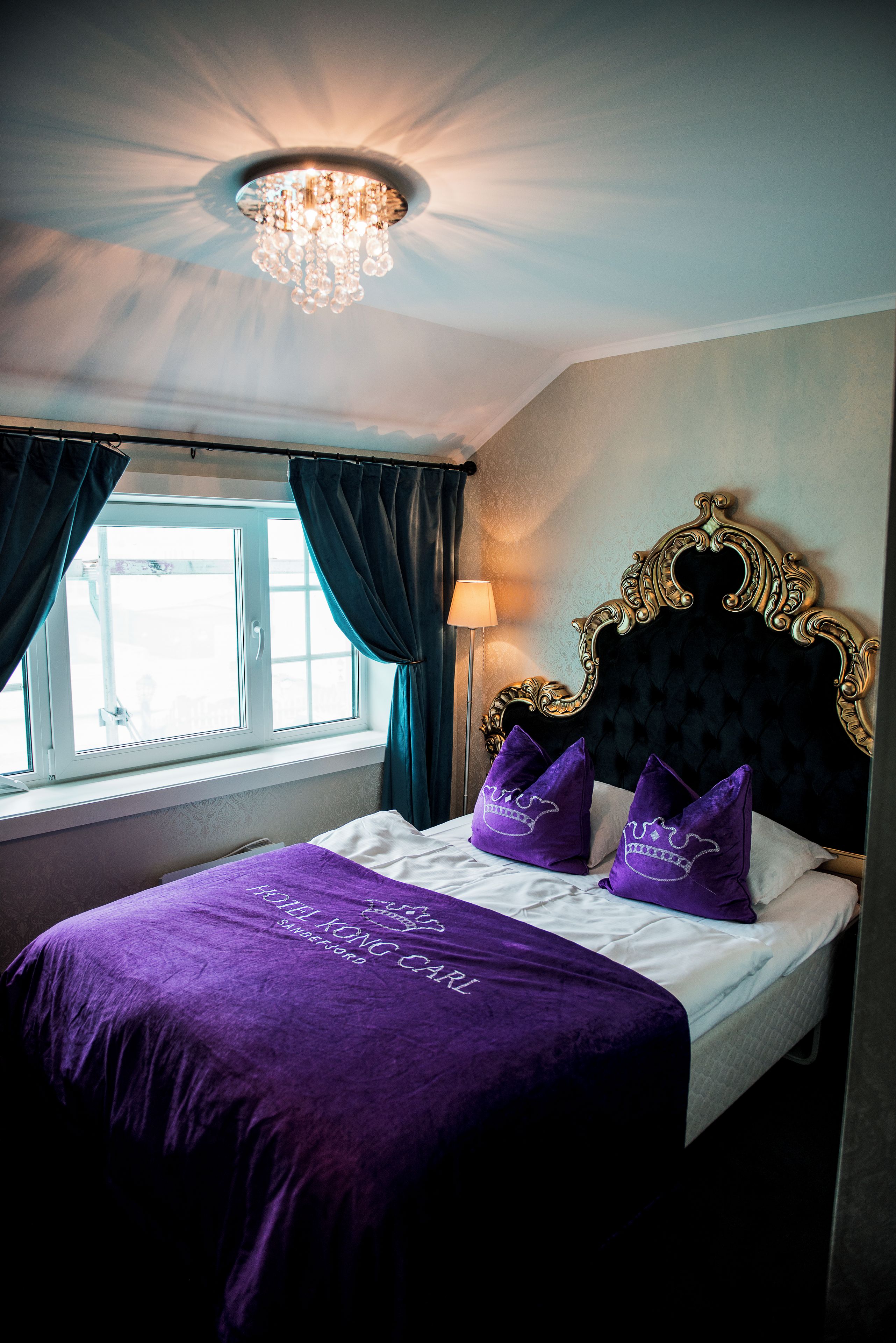 Une chambre d’hôtel avec un lit, un lustre, une fenêtre avec des rideaux et une lampe.