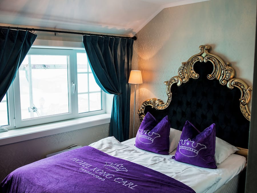 Une chambre d’hôtel avec un lit, un lustre, une fenêtre avec des rideaux et une lampe.
