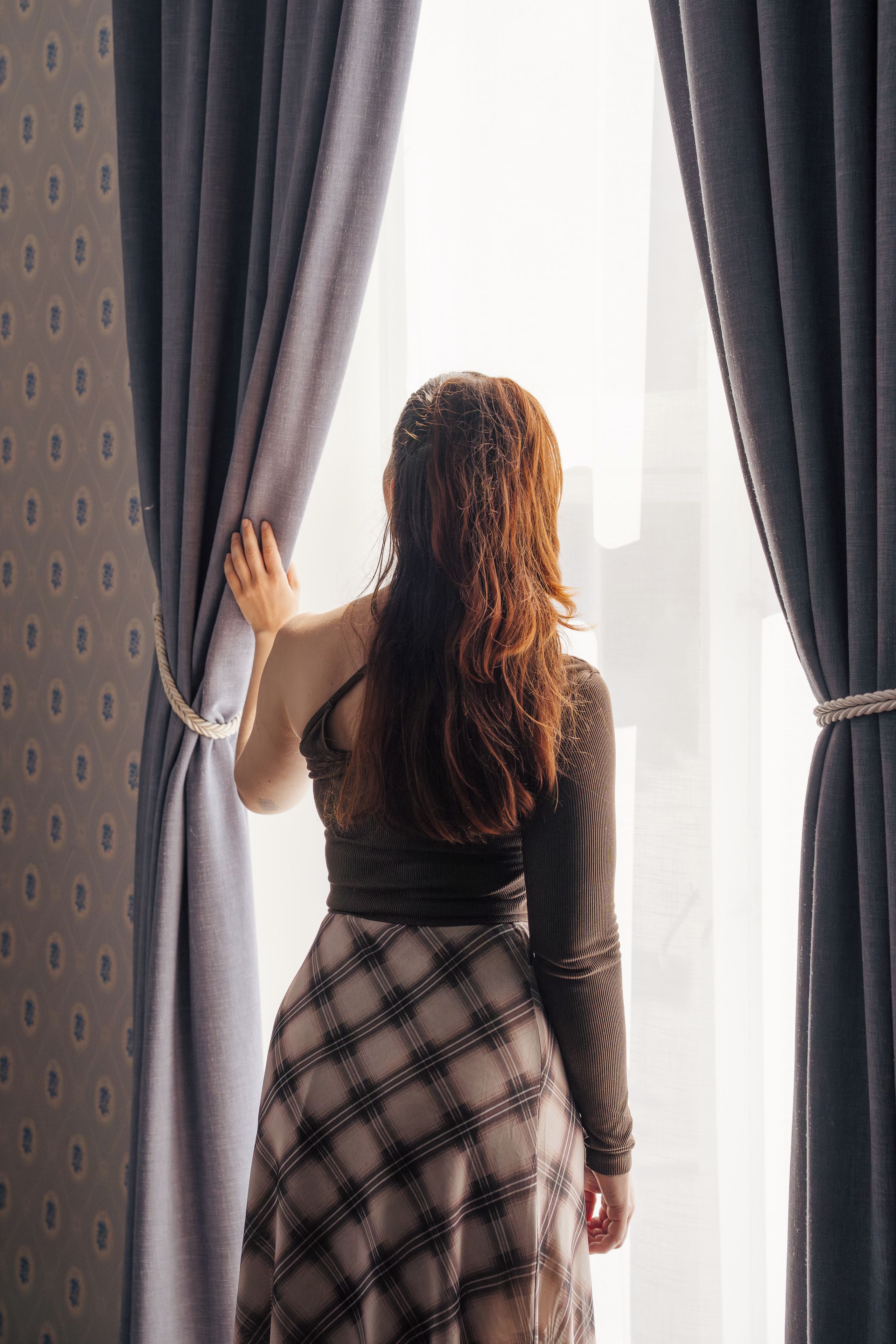 A person standing at a window with curtains, looking outside.