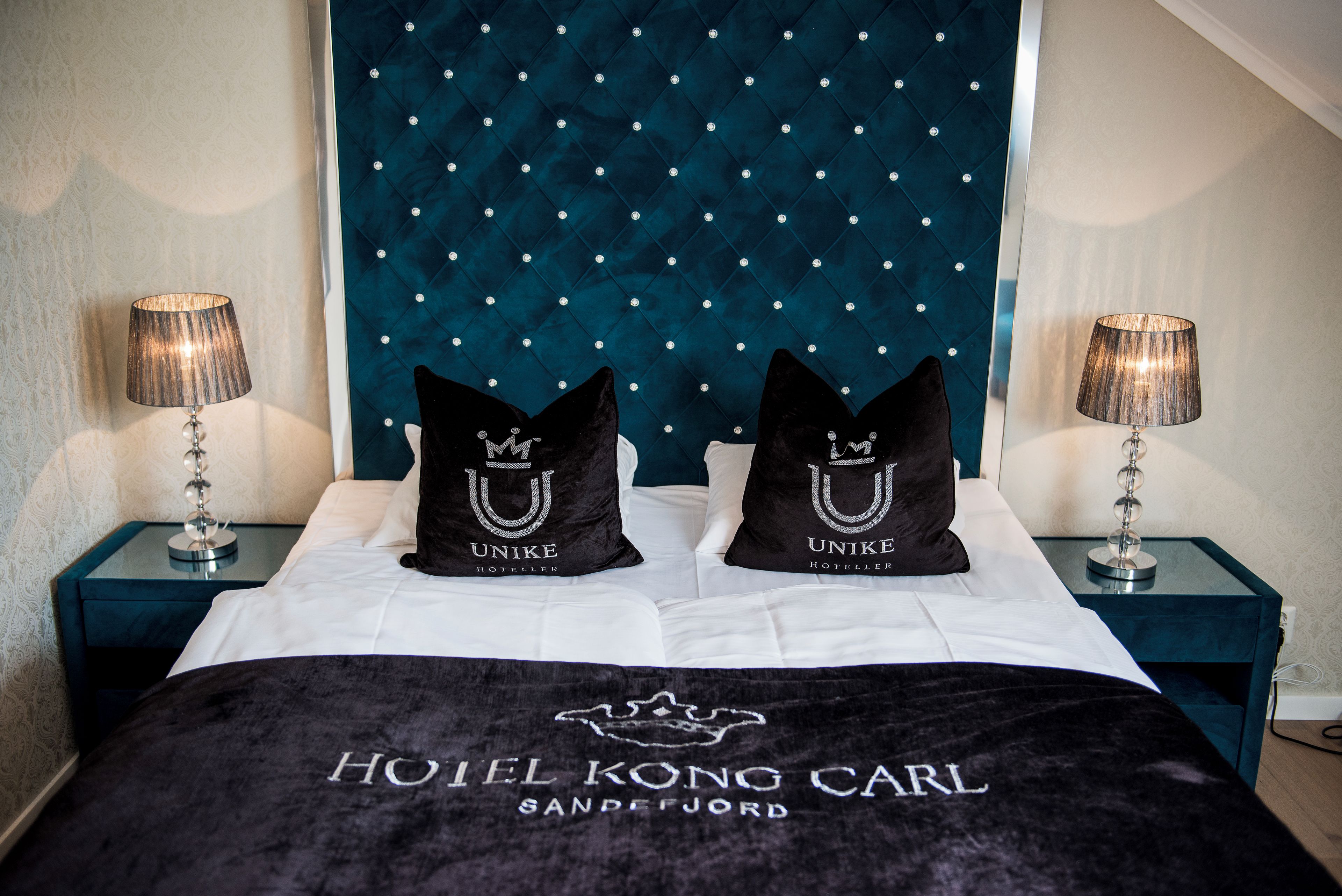 Una habitación de hotel con cabecera capitonada, dos almohadas negras con la marca 'UNIKE HOTEL', una cama con cubierta negra y dos lámparas de noche.