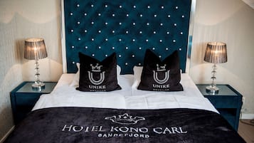 Una habitación de hotel con cabecera capitonada, dos almohadas negras con la marca 'UNIKE HOTEL', una cama con cubierta negra y dos lámparas de noche.