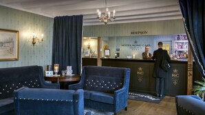 Un vestíbulo de hotel con mostrador de recepción, dos sillones azules, una araña y un cuadro enmarcado en la pared.