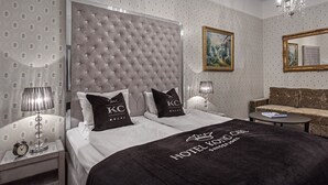 Una habitación de hotel con un cabecero capitonado, dos almohadas negras con la marca "KC", una cama con una colcha negra, una mesa redonda con un libro, un sofá y un candelabro.