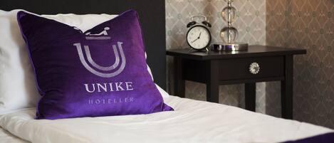 A neatly made bed with a purple pillow featuring the UNIKE HOTELLER logo, a bedside table with an alarm clock, and patterned wallpaper.