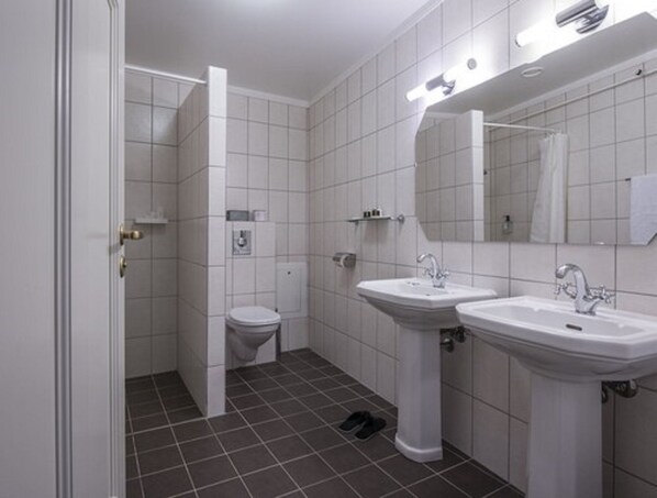 A bathroom with white tiles, a toilet, two sinks, a mirror, and a pair of black slippers on the floor. - Hotel Kong Carl - Unike Hoteller (Sandefjord)