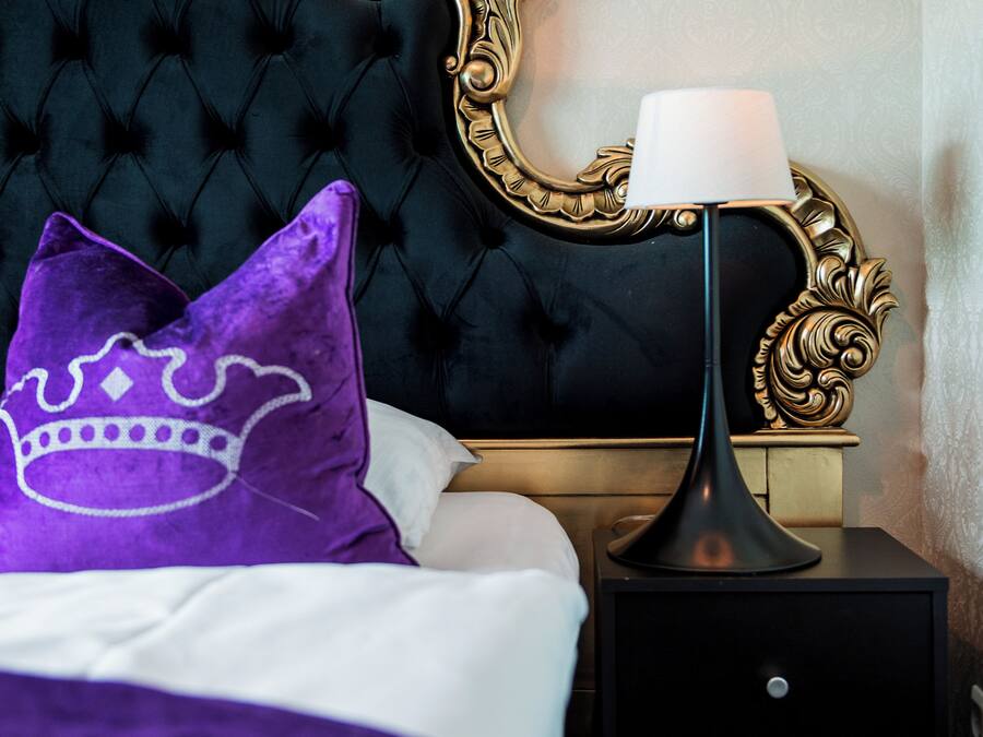 Una cama con cabecera negra, almohada morada, almohada blanca y mesita de noche negra con una lámpara.