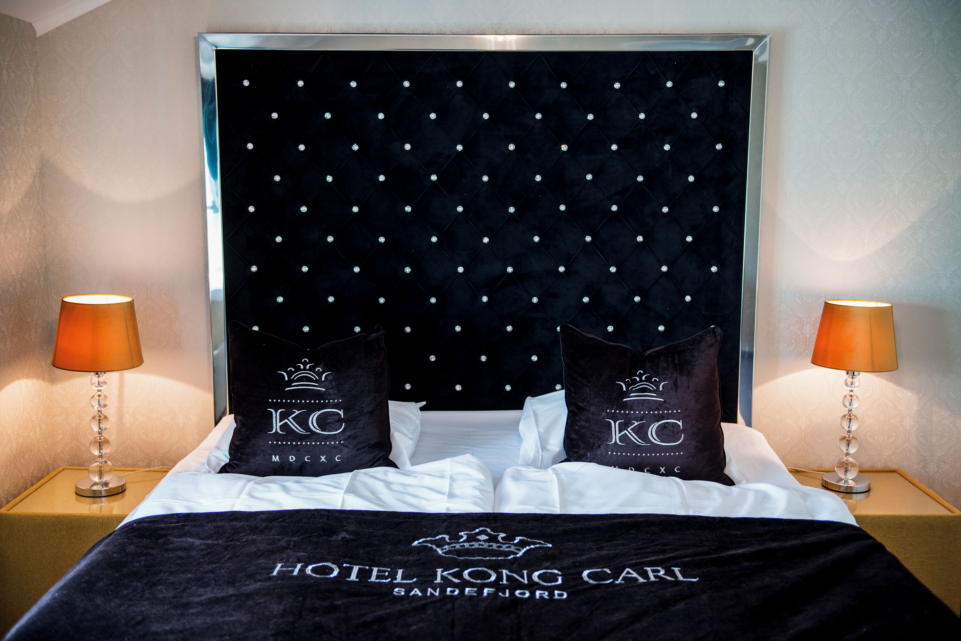 Habitación de hotel con cabecera tapizada en negro, dos almohadas con la marca 'HOTEL KONG CARL', una colcha negra y dos lámparas de noche.