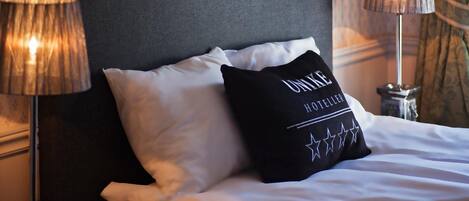 Una cama bien tendida con sábanas blancas y un almohadón negro con texto. Hay dos lámparas a cada lado de la cama.