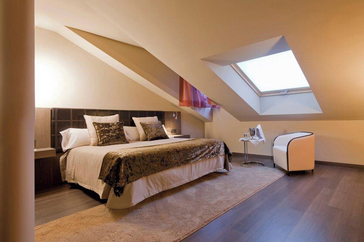 Un dormitorio con techo inclinado, un tragaluz, una cama con cabecera, una mesita de noche, una silla y una pequeña mesa con lámpara.