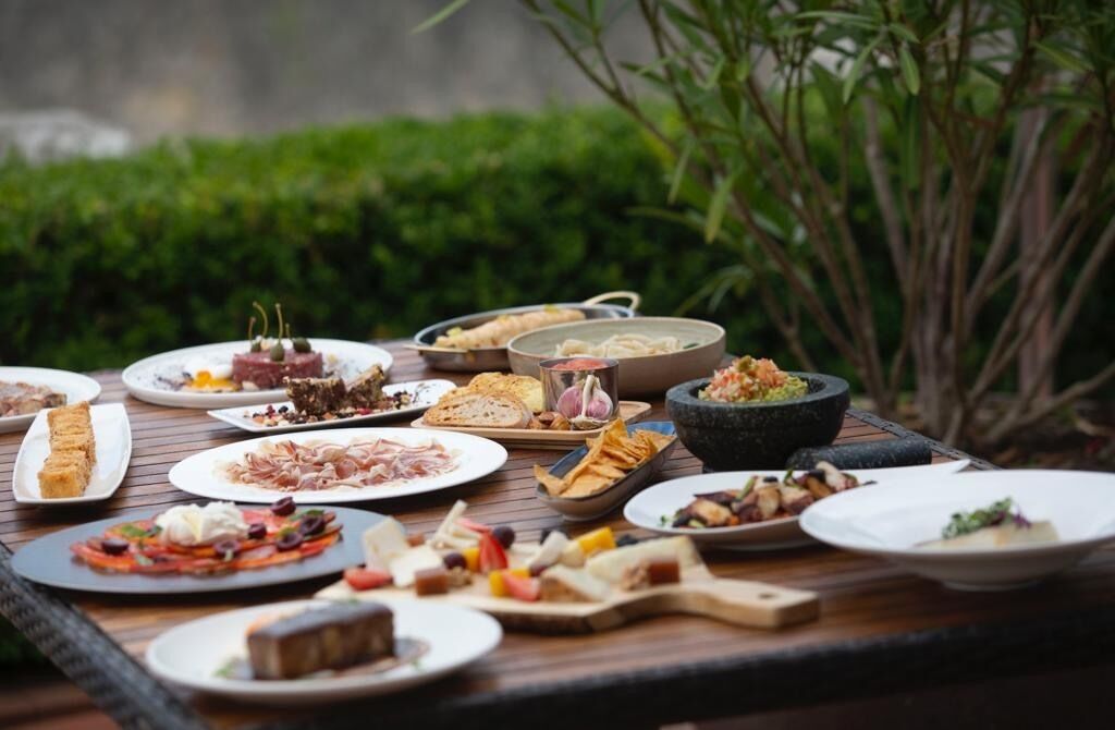 A variety of dishes on a wooden table outdoors with greenery in the background.