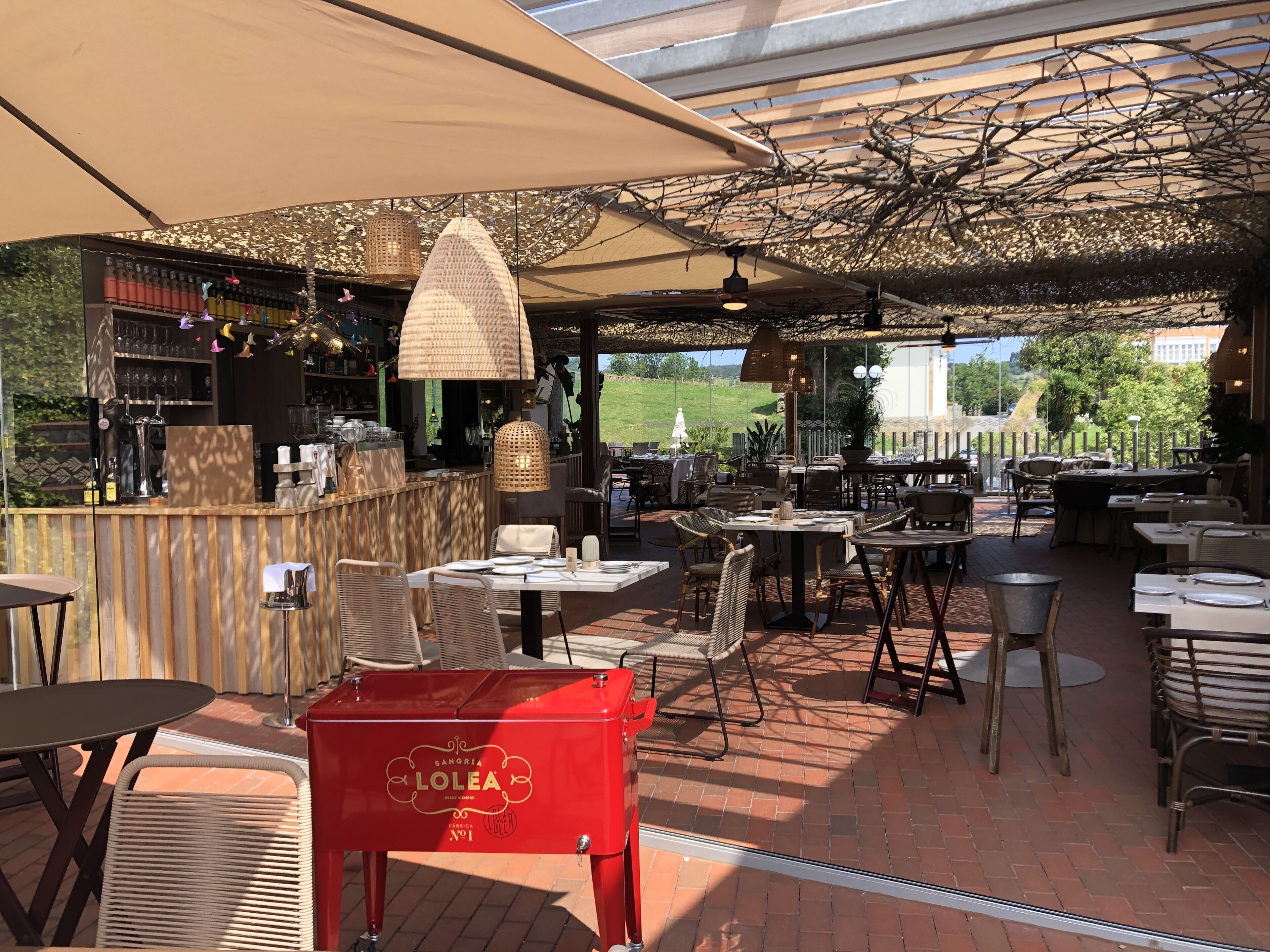 Een terras buiten met tafels en stoelen, een bar en een rode grill met de merknaam 'LOLEA'.