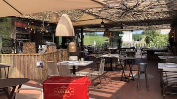 Een terras buiten met tafels en stoelen, een bar en een rode grill met de merknaam 'LOLEA'.