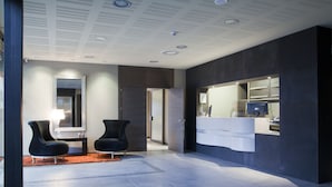 A modern hotel lobby with a reception desk, two black armchairs, a large mirror, and a dark-colored wall with a built-in counter. - La Piconera And Spa (Ribadesella)