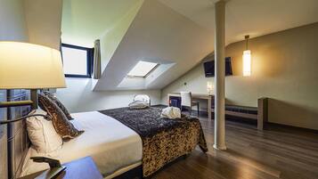 A modern hotel room with a sloped ceiling, a bed with a black and white patterned bedspread, a desk with a chair, a TV, and a lamp.