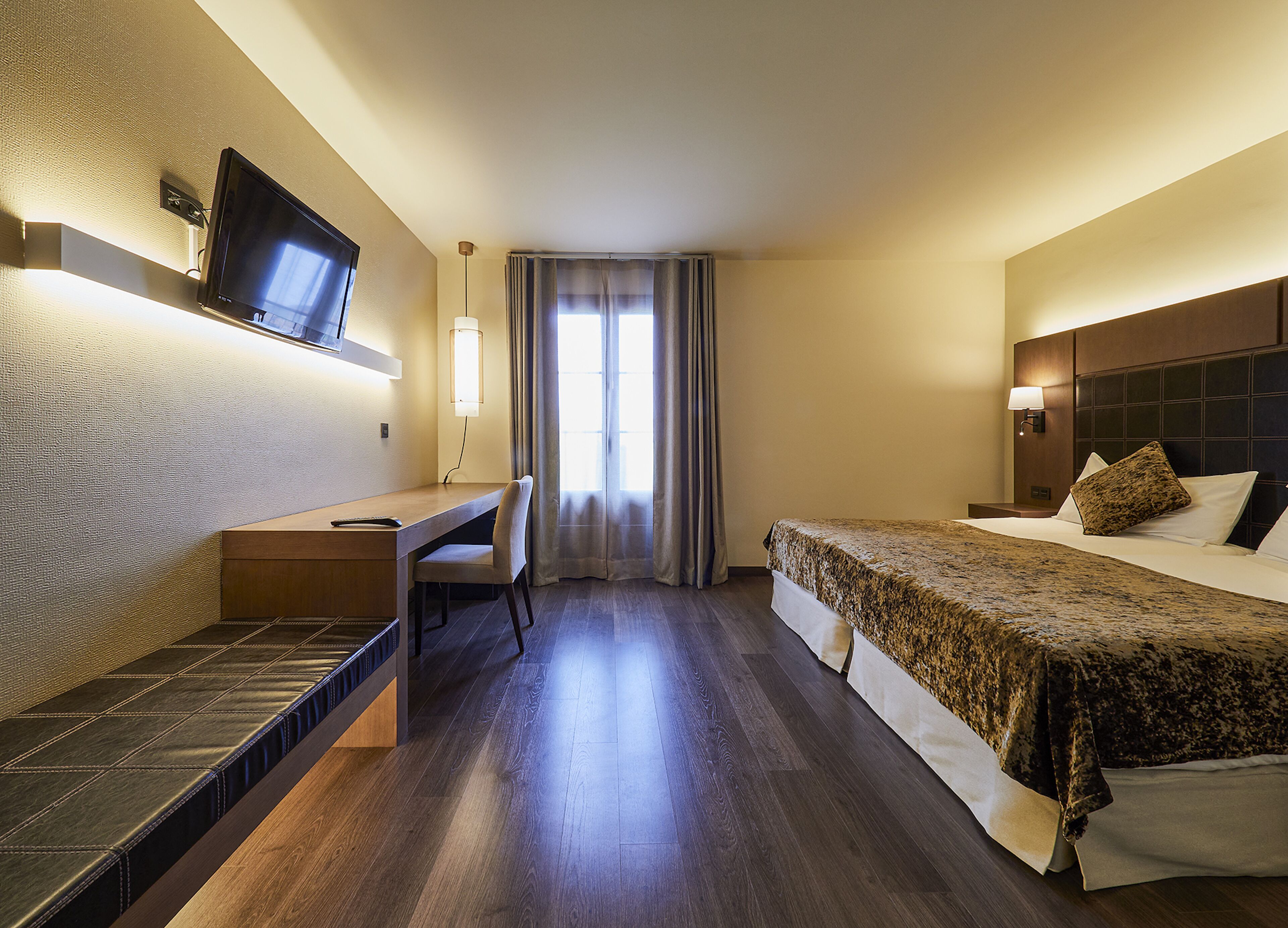 A hotel room with a bed, a desk, a chair, a television mounted on the wall, and a window with curtains.