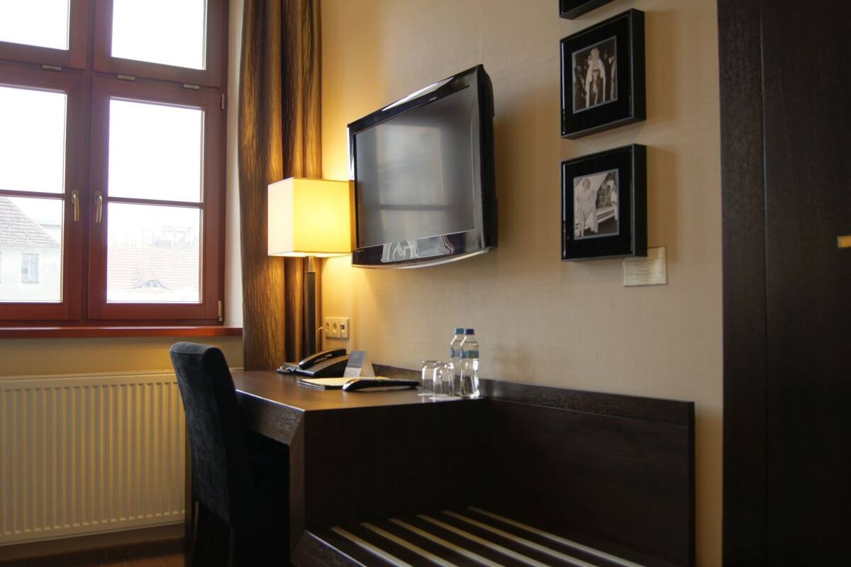 A hotel room with a desk, a television mounted on the wall, a lamp, a chair, and framed pictures.
