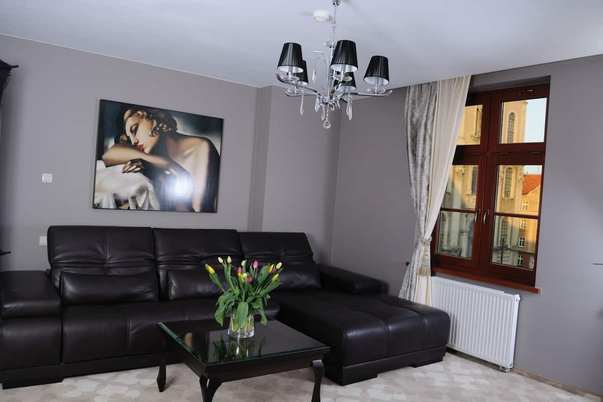 A living room with a black leather sofa, a glass coffee table with a vase of flowers, a painting on the wall, and a window with curtains.