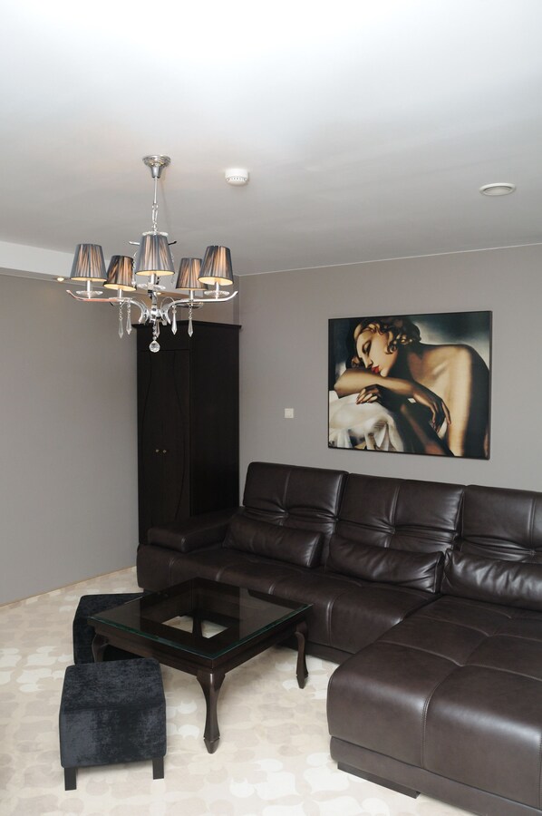 A living room with a dark brown leather sofa, a glass coffee table, a black ottoman, a chandelier, and a painting on the wall.