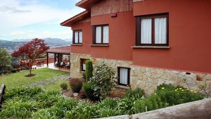 Una casa roja de dos pisos con base de piedra, múltiples ventanas y un jardín con diversas plantas y flores.
