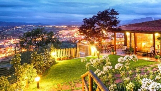 Vista de un paisaje urbano al anochecer, con un área de estar exterior iluminada y un jardín lleno de flores.